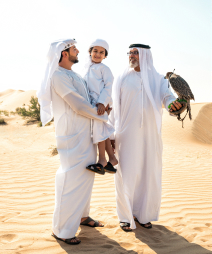 Мужчина держит на руках своего сына, стоя рядом с отцом, у которого на руке орел; они улыбаются, вокруг пустыня.