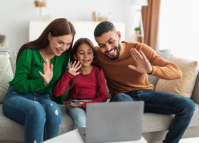 Улыбающаяся семья машет рукой в сторону экрана ноутбука во время видеозвонка.