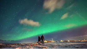 Photo of a couple standing in a field illuminated by the Northern Lights.